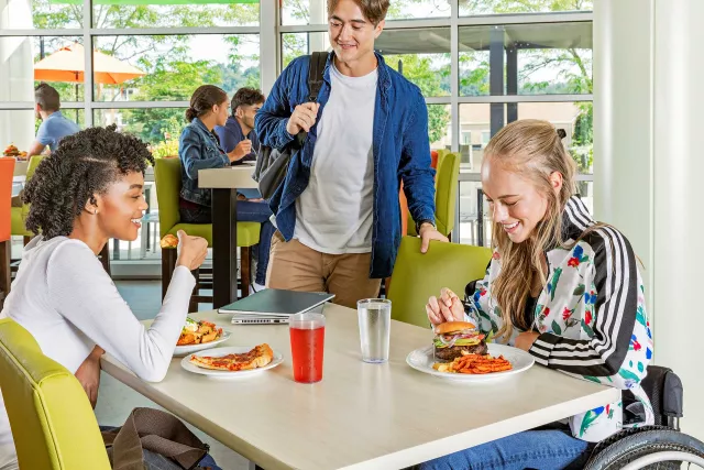 Two college students eating in cafeteria while another comes to join them